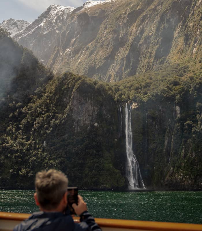 Person taking photo of waterfall cascading down mountains next to a green lake
