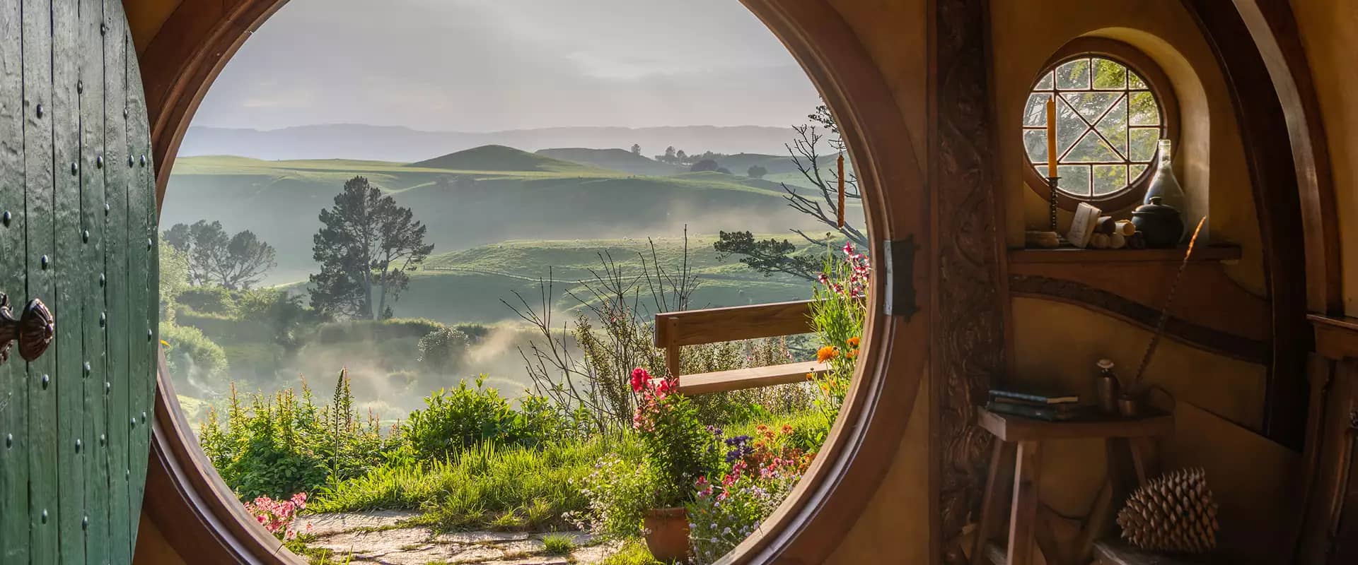 Inside view of a hobbit hole with a round door open to a green garden and rolling hills beyond.